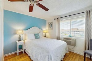 A bedroom with a bed, two bedside lamps, a ceiling fan, a window with curtains, and a view of the outside. - Coastal Tranquility Retreat (North Myrtle Beach)