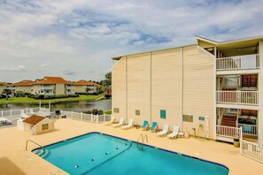 Ein Schwimmbecken mit Liegestühlen und ein kleines Gebäude am Pool, neben einem mehrstöckigen Gebäude mit Balkonen.
