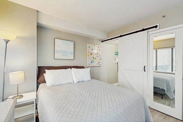 Quarto com cama, mesa de cabeceira, abajur, porta e parede com um quadro.