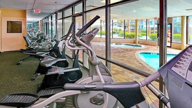 Ett gym med löpband och motionscyklar, stora fönster och utsikt över en utomhuspool.