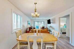 A dining area with a wooden table and white chairs, a chandelier, and a view of a living room with a sofa and a television. - Shipwatch Pointe II P-105 (Myrtle Beach)
