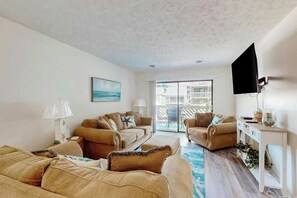 A living room with two sofas, a coffee table, a TV, and a sliding glass door leading to an outdoor area. - Shipwatch Pointe II P-105 (Myrtle Beach)