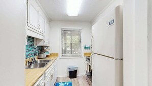 Een kleine keuken met witte kastjes, een roestvrijstalen spoelbak en een raam met zonwering.