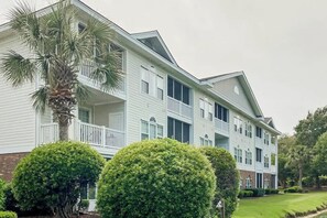 A multi-story building with balconies, a well-maintained lawn, and mature trees. - River Crossing Unit 131 (North Myrtle Beach)