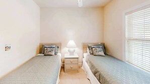 A bedroom with two single beds, a ceiling fan, a bedside table with a lamp, and a window with blinds. - River Crossing Unit 131 (North Myrtle Beach)