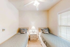A bedroom with two single beds, a ceiling fan, a bedside table with a lamp, and a window with blinds. - River Crossing Unit 131 (North Myrtle Beach)