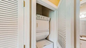 A white laundry room with a built-in washer and dryer, a door with a brass handle, and a vent above the dryer. - River Crossing Unit 131 (North Myrtle Beach)
