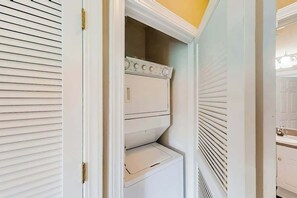 A white laundry room with a built-in washer and dryer, a door with a brass handle, and a vent above the dryer. - River Crossing Unit 131 (North Myrtle Beach)