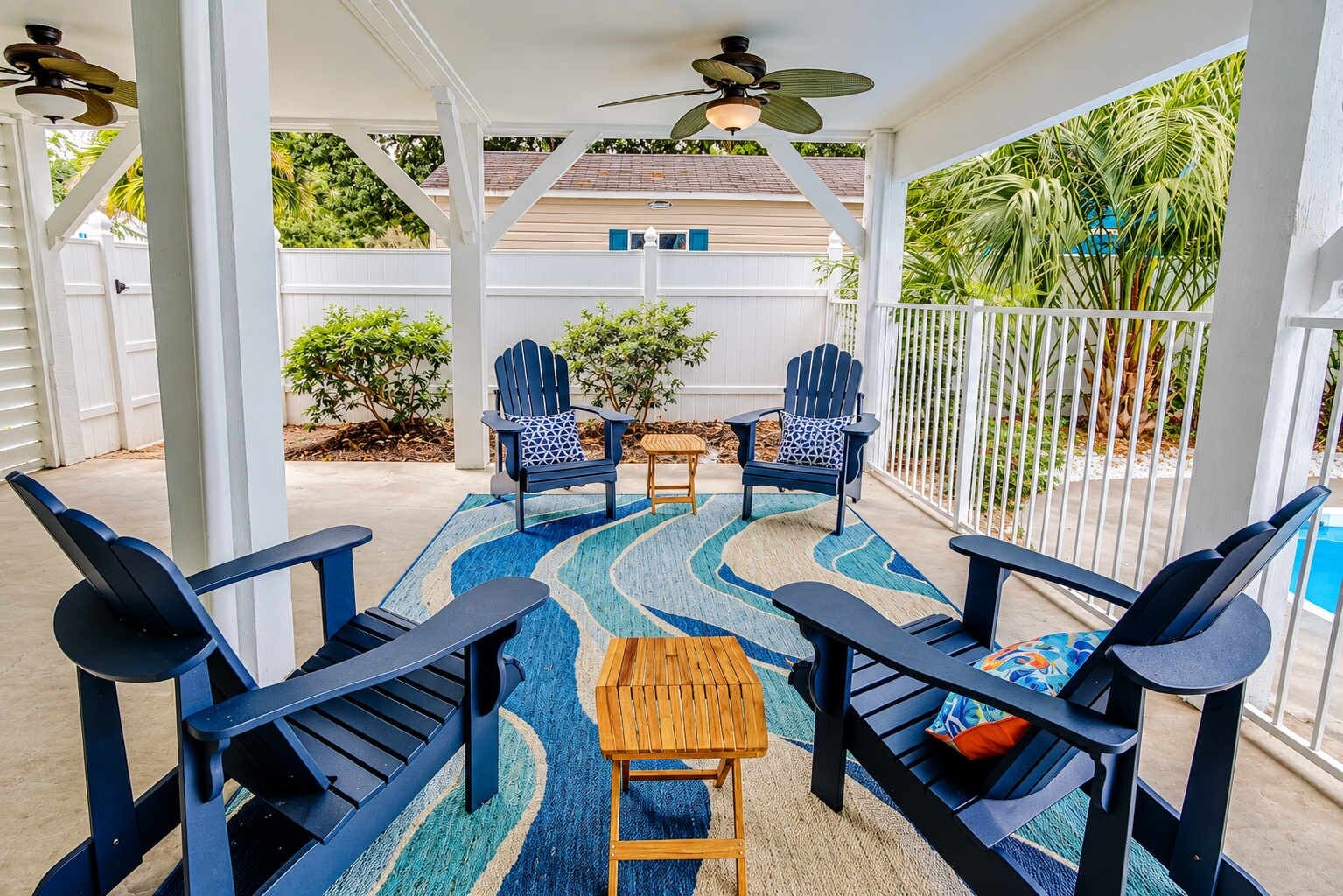 An outdoor patio with Adirondack chairs, a wooden table, and a patterned rug.