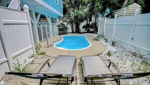 A backyard with a swimming pool, two lounge chairs, a white fence, and a blue house. - Surfside Escape Retreat (Surfside Beach)