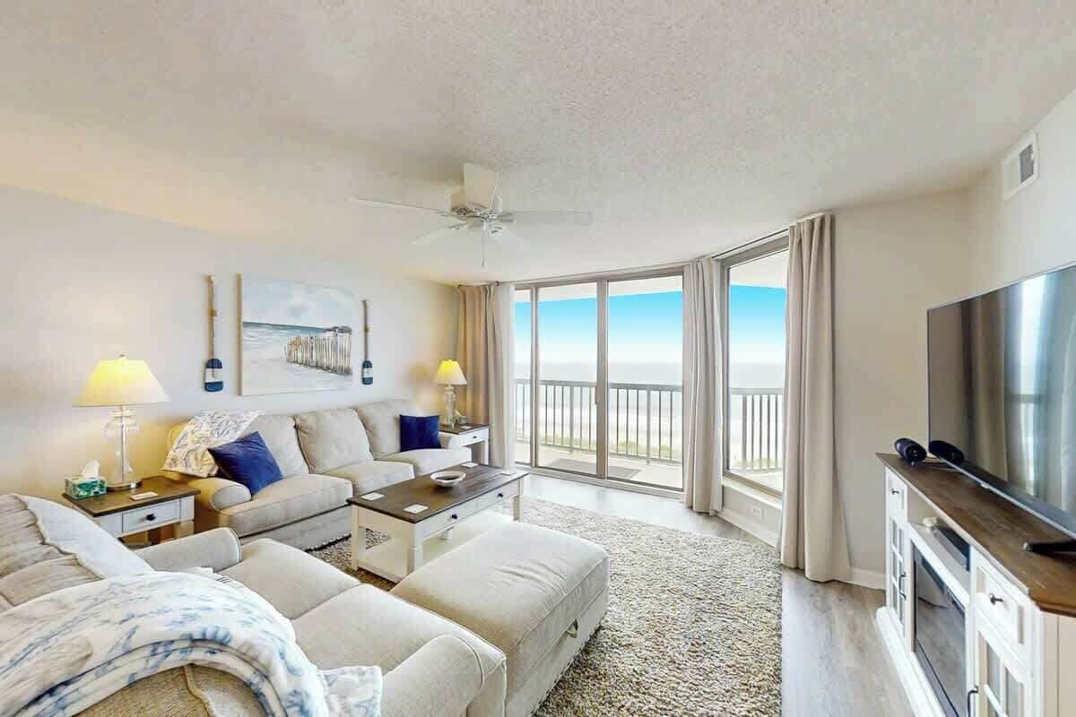 A hotel room with a balcony, a flat-screen TV, a sofa, a coffee table, and a ceiling fan.
