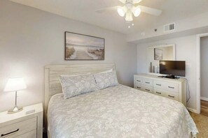 Une chambre à coucher comprenant un lit, une table de chevet, une lampe, un ventilateur de plafond, un téléviseur et une commode.