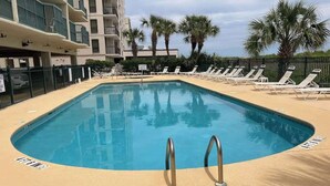A rectangular pool with two metal ladders, surrounded by lounge chairs and palm trees, with a multi-story building in the background. - Endless Summer Oasis (North Myrtle Beach)