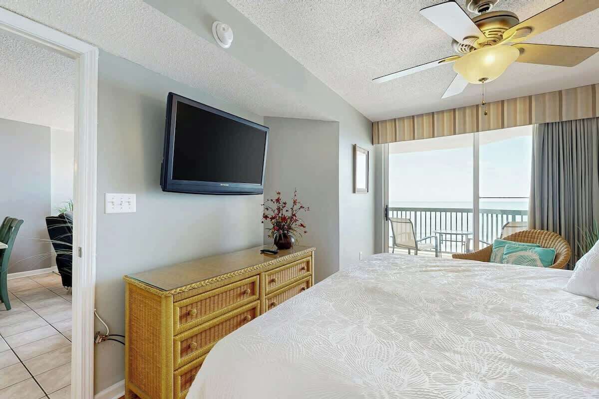 Habitación de hotel con una cama, televisor montado en la pared, ventilador de techo, ventana con cortinas y balcón con sillas.