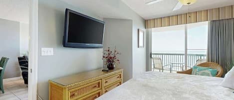 A hotel room with a bed, a television mounted on the wall, a ceiling fan, a window with curtains, and a balcony with chairs.