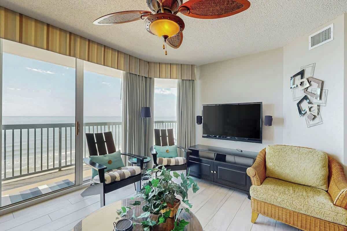 A modern living room with a large TV, comfortable seating, a balcony with a view, and a ceiling fan.