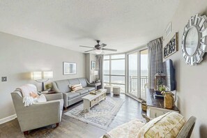 A spacious living room with a ceiling fan, a large window with curtains, a sofa, a coffee table, and a television. - Sunrise Tides at the Cherry Grove Pier (North Myrtle Beach)