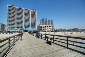 A wooden boardwalk leading to a beach with high-rise buildings in the background. - Sunrise Tides at the Cherry Grove Pier (North Myrtle Beach)