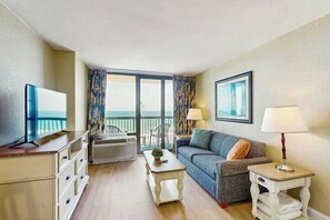 Ein Wohnzimmer mit Fernseher, einer Couch, einem Couchtisch und einer Lampe. Es gibt einen Balkon mit Aussicht.