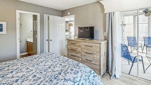 Ein Schlafzimmer mit einem Bett, einem Schminktisch, einem Fernseher und einer Glastür, die auf einen Balkon mit Stühlen führt.