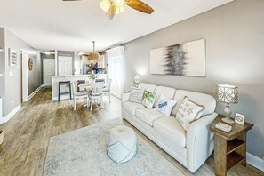 A modern living room with a white sofa, a round ottoman, and a dining area with a white table and chairs. - Waterway Landing 216-A (North Myrtle Beach)