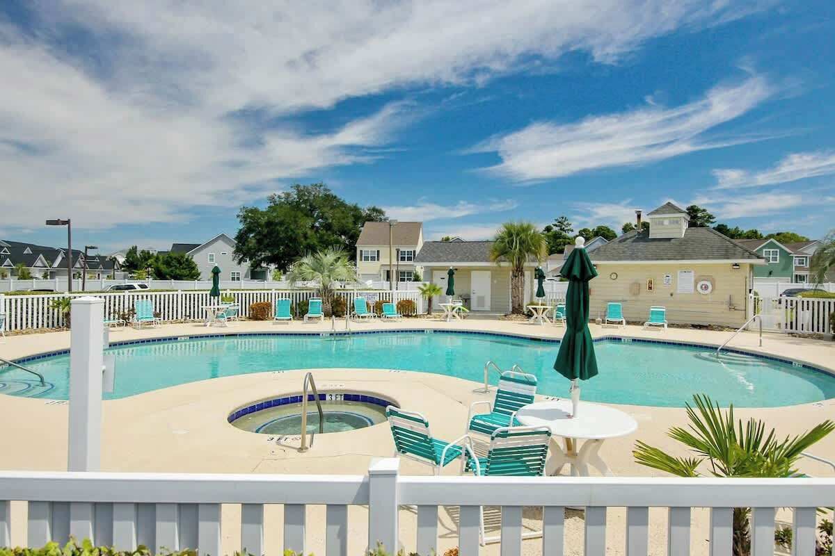 En swimmingpool med solstolar och parasoller, omgiven av ett vitt staket och med byggnader i bakgrunden.