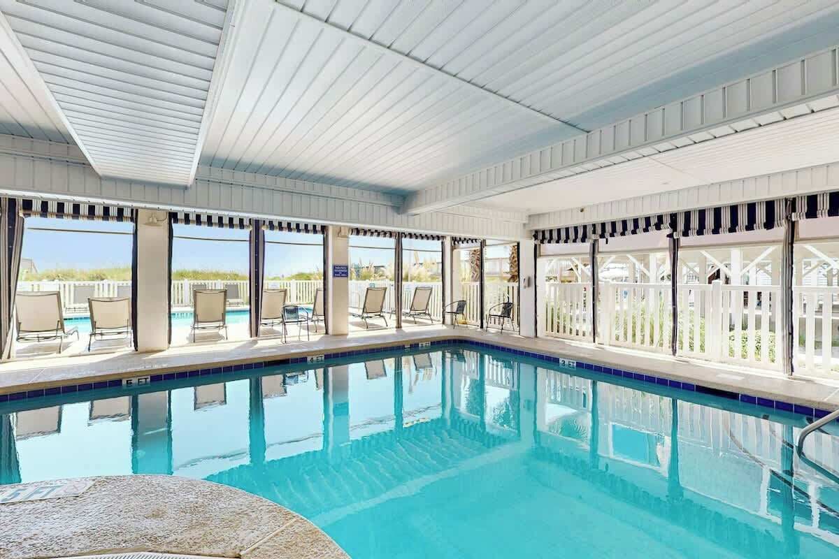Una piscina con agua azul cristalina, rodeada de sillones de descanso y una zona cubierta con amplios ventanales.