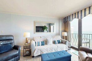 A hotel room with a sofa, a chair, a small table with a lamp, a window with curtains, and a balcony with a railing. - Coastal Living Retreat (Myrtle Beach)