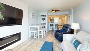 Ein Wohnzimmer mit Flachbildfernseher, Kamin, Essbereich mit Tisch und Stühlen, einer Küche im Hintergrund und einem blauen Sofa mit Lampe.