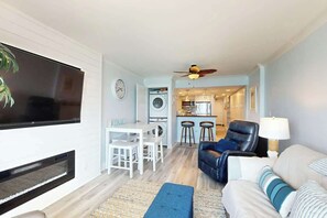 Ein Wohnzimmer mit Flachbildfernseher, Kamin, Essbereich mit Tisch und Stühlen, einer Küche im Hintergrund und einem blauen Sofa mit Lampe.