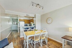 A modern kitchen with white cabinets, a wooden dining table, and a clock on the wall. - Tilghman Beach & Racquet 259 (North Myrtle Beach)