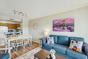 A modern living room with a blue sofa, dining table, and chairs. There is a painting of a Ferris wheel on the wall. - Tilghman Beach & Racquet 259 (North Myrtle Beach)