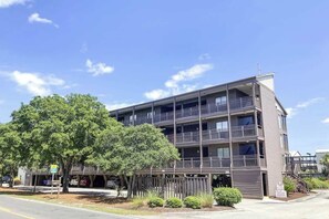Un immeuble à plusieurs étages doté de balcons, d’un espace de stationnement et d’un arbre devant.