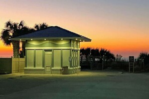 Ein kleines, freistehendes Gebäude mit flachem Dach, vermutlich ein Strandhaus oder eine kleine Ferienanlage, mit einer einzigen Tür und einem kleinen Fenster, umgeben von Palmen und einem Sonnenuntergang im Hintergrund.