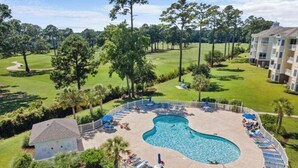 A pool area with lounge chairs and a small building, surrounded by a fence and trees. - Luster Leaf Unit 303 (Myrtle Beach)