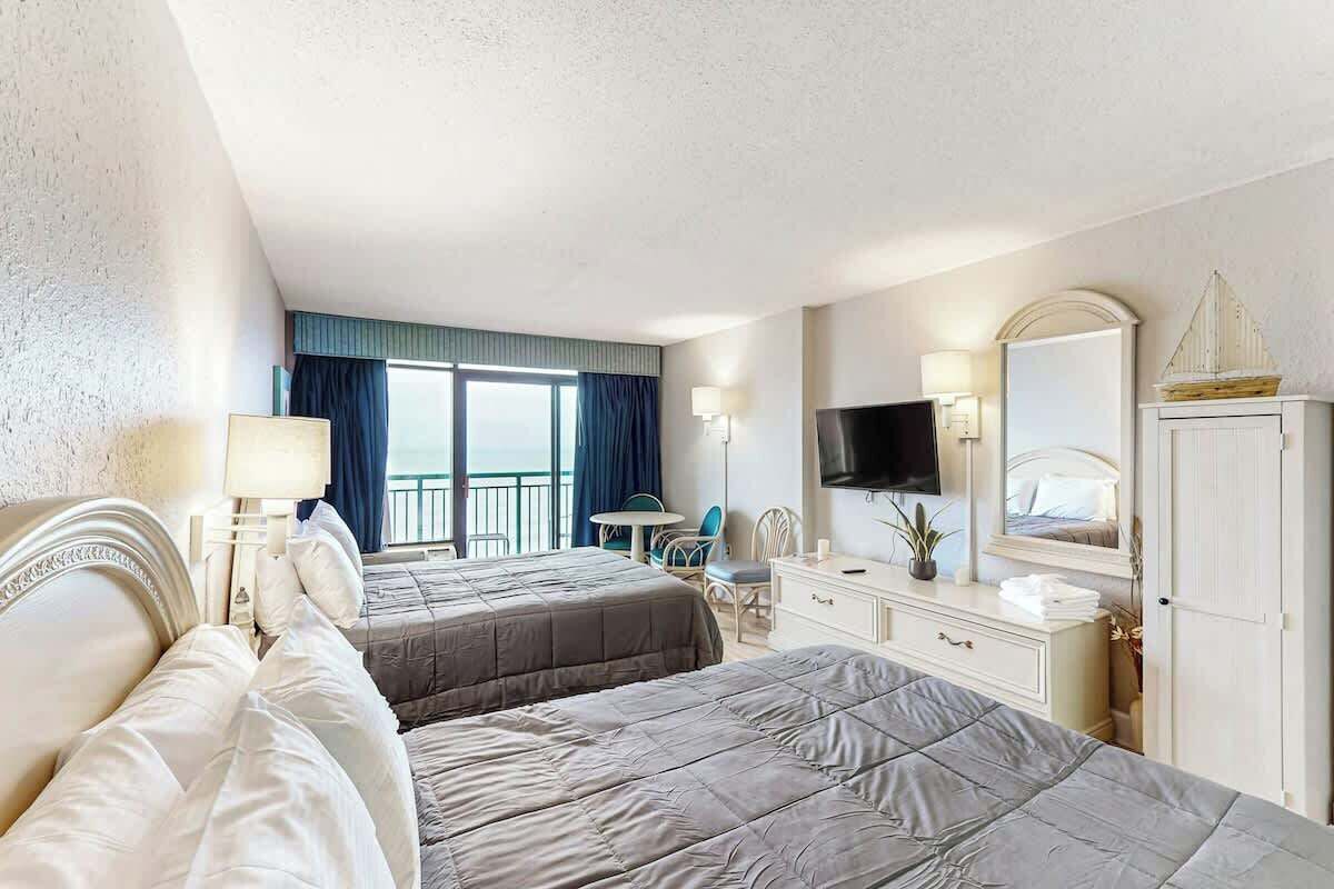A hotel room with two beds, a TV, a desk, and a balcony with a view.
