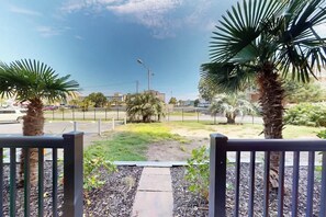 A gated entrance with a pathway leading to a landscaped area with palm trees and a view of a street with buildings and parked cars. - Holiday Towers Unit 104 (Myrtle Beach)