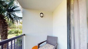 A balcony with a wicker chair, a small orange stool, and a palm tree. - Holiday Towers Unit 104 (Myrtle Beach)
