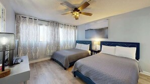A hotel room with two beds, a ceiling fan, a desk with a lamp, and a window with curtains. - Holiday Towers Unit 104 (Myrtle Beach)
