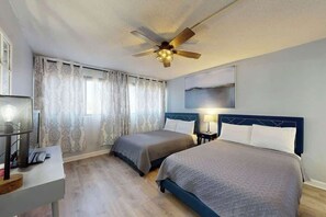 Ein Hotelzimmer mit zwei Betten, einem Deckenventilator, einem Schreibtisch mit Lampe und einem Vorhangfenster.