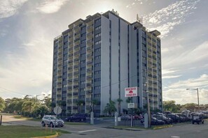 A multi-story building with balconies, a sign indicating 'Holiday Towers,' and a parking lot with cars. - Holiday Towers Unit 104 (Myrtle Beach)