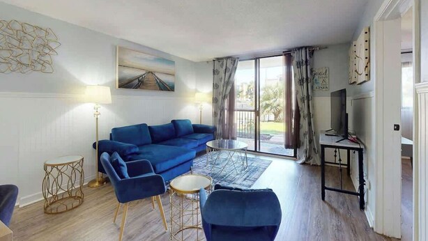 A living room with a blue sofa, a coffee table, and a lamp. There is a painting on the wall and a window with curtains. - Holiday Towers Unit 104 (Myrtle Beach)
