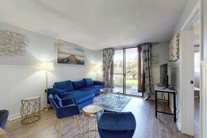 Een woonkamer met een blauwe bank, een salontafel en een lamp. Aan de muur hangt een schilderij en er is een raam met gordijnen.