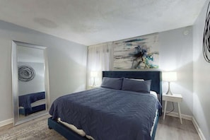 A bedroom with a bed, a nightstand, a mirror, and a painting on the wall. - Holiday Towers Unit 104 (Myrtle Beach)