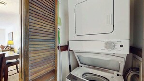 A compact laundry area with a front-loading washing machine and dryer, a wooden door, and a dining area with a table and chairs. - Shipwatch Pointe II  P-302 (Myrtle Beach)