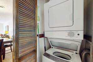 A compact laundry area with a front-loading washing machine and dryer, a wooden door, and a dining area with a table and chairs. - Shipwatch Pointe II  P-302 (Myrtle Beach)
