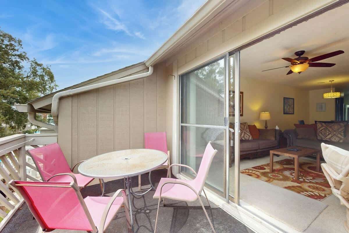 A balcony with a table and chairs, a sliding glass door leading to a living room with a sofa and a ceiling fan.