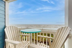 Ein Balkon mit zwei Adirondack-Stühlen und einem kleinen runden Tisch, der Blick auf einen Strand bietet.
