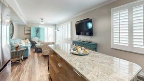 A modern kitchen with a marble countertop, a built-in TV, and a dining area with a blue sofa and wicker chairs. - Summer Place Unit B-4 (North Myrtle Beach)