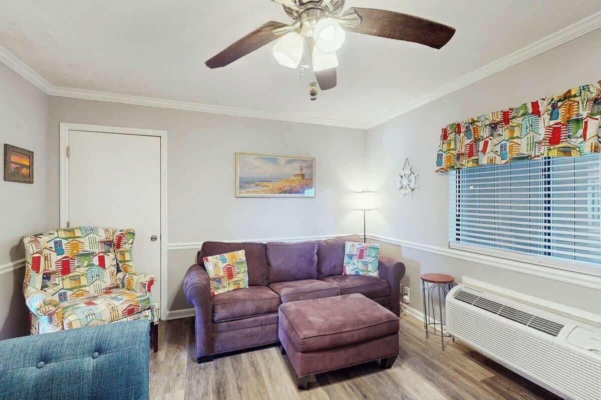 A living room with a ceiling fan, a sofa, an ottoman, a patterned armchair, a painting, and a window with blinds.